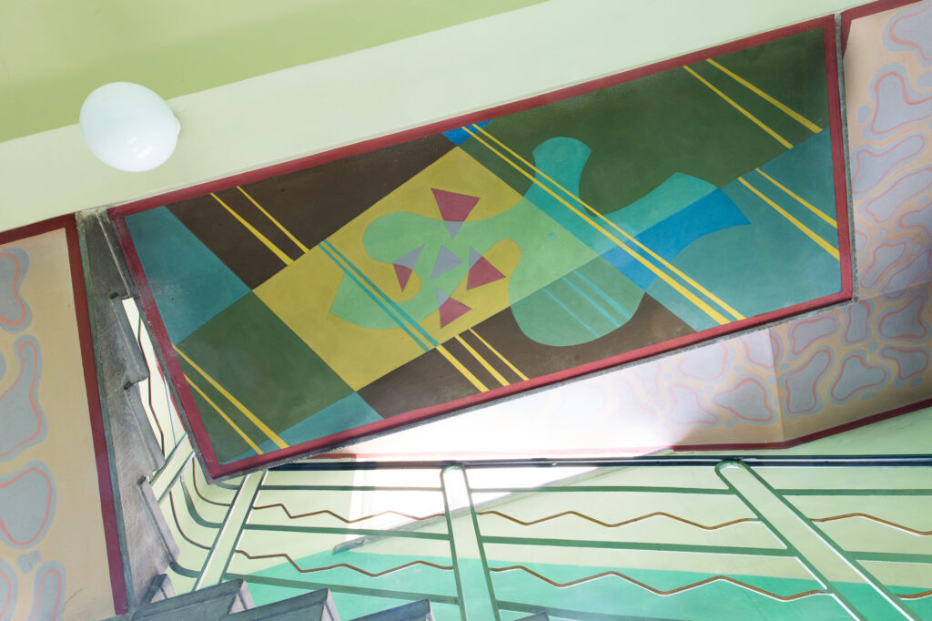 Geometryczne malarstwo sufitowe nad biegiem schodów w modernistycznym wnętrzu.