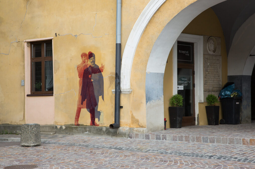 Dwuwarstwowy kolaż w czerwieni i błękicie przedstawiający historyczną postać na elewacji kamienicy w Jarosławiu.